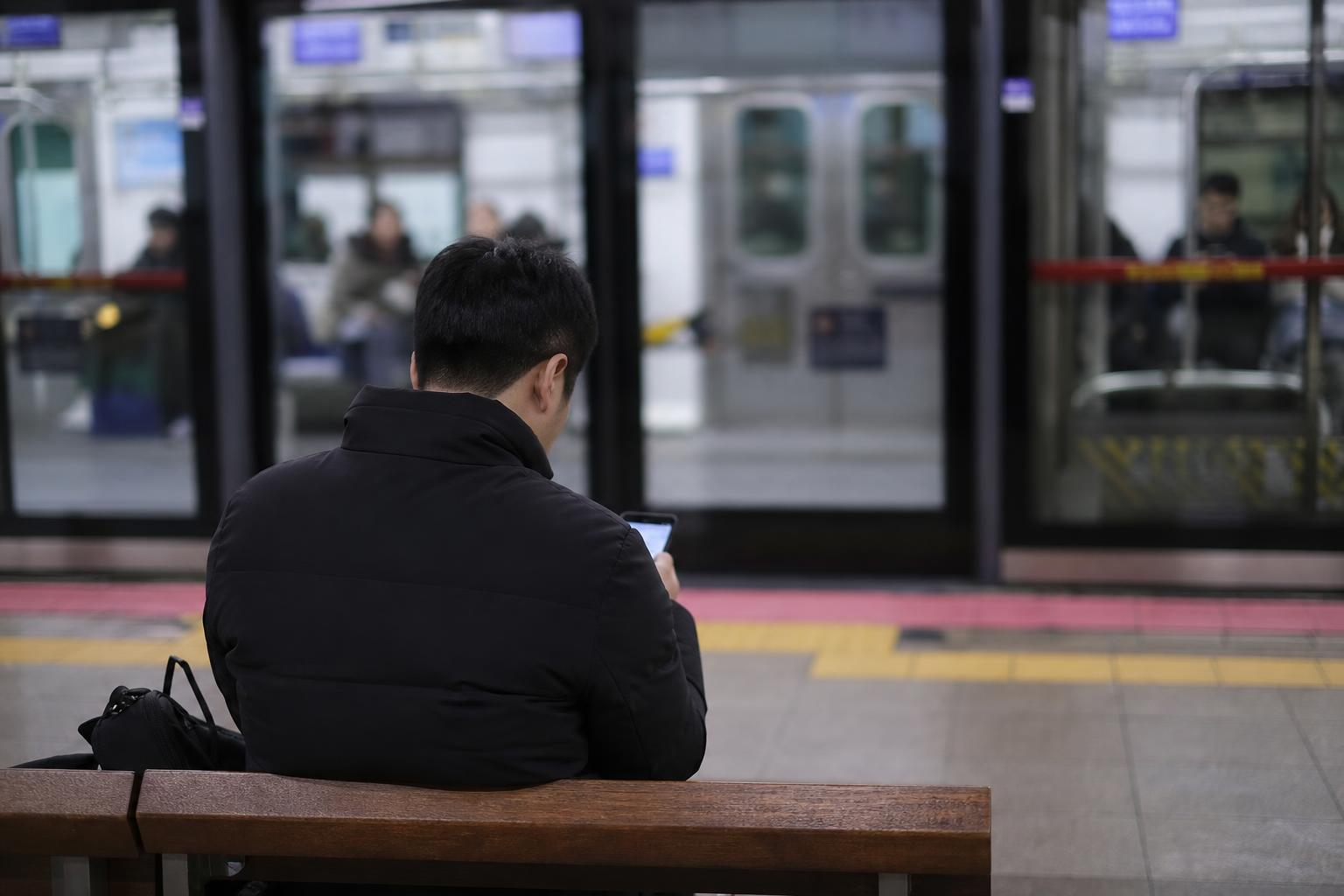 지하철 승강장 의자에 앉아 스마트폰을 들여다보는 사람의 뒷모습